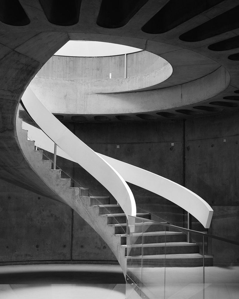 A sculptural spiral staircase rising through a concrete atrium toward an oval skylight